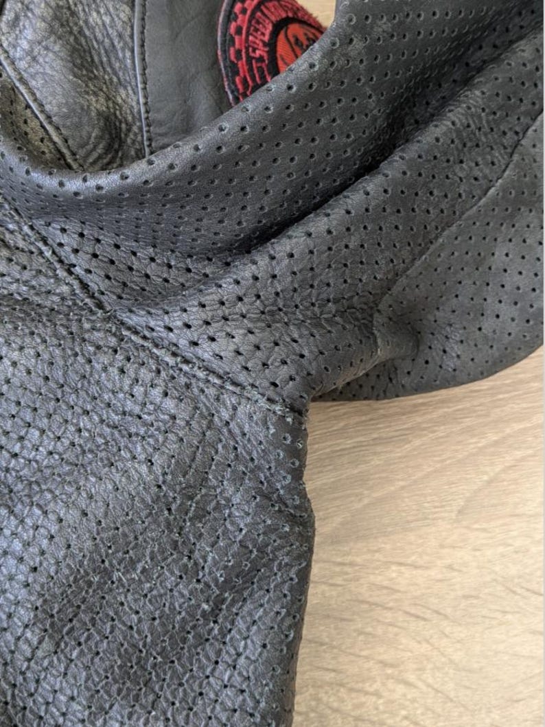 May include: Close-up of a black leather jacket with perforated detailing. The jacket has a patch with red and white text. The leather appears textured and the jacket is folded.