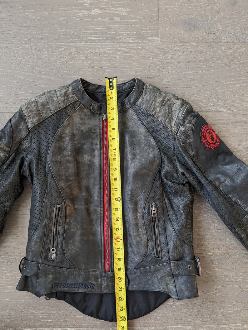 May include: A distressed black leather motorcycle jacket with a red zipper and the text "SPEED AND STRENGTH". The jacket has a round collar, zippered pockets, and a patch on the sleeve. The jacket measures approximately 24 inches in length.
