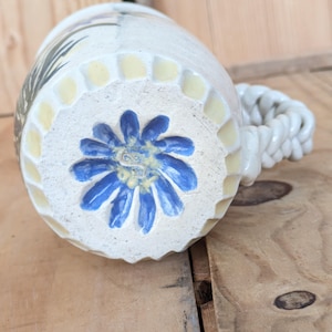 Puede incluir: Taza de cerámica blanca con un diseño de flor azul y amarilla en la parte inferior. La taza tiene un asa trenzada.