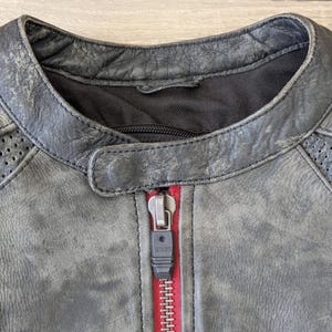 May include: Close-up of a distressed gray leather jacket with a red zipper. The jacket features a round collar, quilted shoulder details, and perforated panels. The zipper is a silver color.