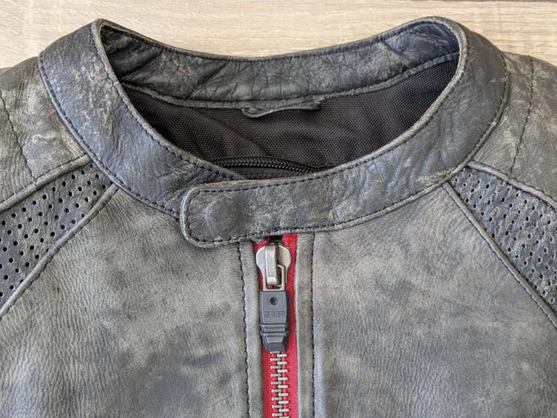 May include: Close-up of a distressed gray leather jacket with a red zipper. The jacket features a round collar, quilted shoulder details, and perforated panels. The zipper is a silver color.