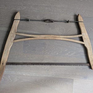 Puede incluir: Una sierra de arco antigua, con un marco de madera y una hoja de acero dentada. El marco de madera clara tiene una forma curvada. La hoja de sierra es de color gris oscuro, y el mecanismo de tensión es de metal. Una herramienta clásica de carpintería.