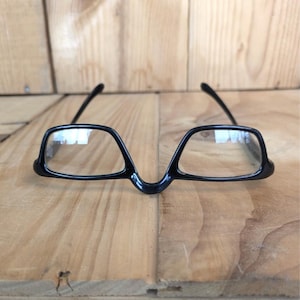 Puede incluir: Gafas con montura negra de diseño angular único. Las gafas presentan un puente distintivo y lentes rectangulares. Las patillas son finas y se extienden hacia atrás desde la montura. Las gafas están sobre un fondo de madera.