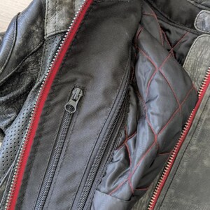 May include: Close-up of a distressed black leather motorcycle jacket. The jacket features a red zipper, quilted lining with red stitching, and multiple zippered pockets. The leather has a worn, textured appearance.