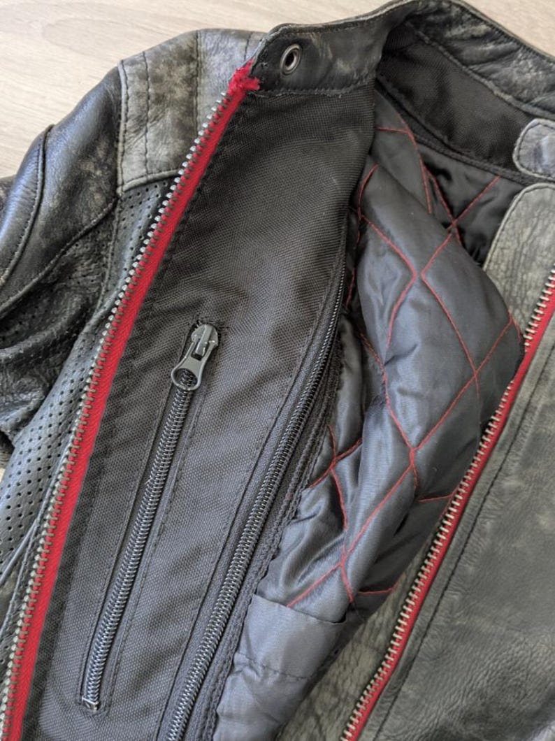 May include: Close-up of a distressed black leather motorcycle jacket. The jacket features a red zipper, quilted lining with red stitching, and multiple zippered pockets. The leather has a worn, textured appearance.
