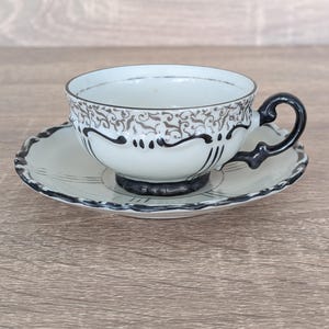 May include: A vintage teacup and saucer set. The cup and saucer are white with black and silver decorative patterns. The cup handle and the rim of the saucer are black. The set is placed on a wooden surface.