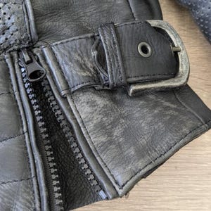 May include: Close-up of a black leather glove with a silver buckle and zipper. The glove has a quilted pattern and perforated detailing. The leather appears worn, with visible texture and slight color variations.
