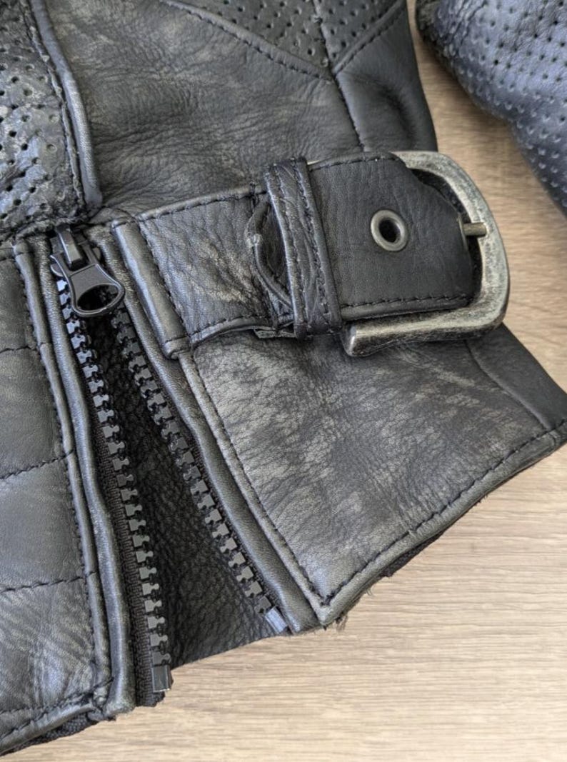 May include: Close-up of a black leather glove with a silver buckle and zipper. The glove has a quilted pattern and perforated detailing. The leather appears worn, with visible texture and slight color variations.