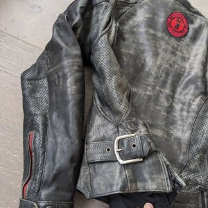 May include: Distressed black leather motorcycle jacket with a red and black patch on the back. The jacket features a buckle closure, perforated sleeves, and red zipper accents. The leather has a worn, vintage appearance.