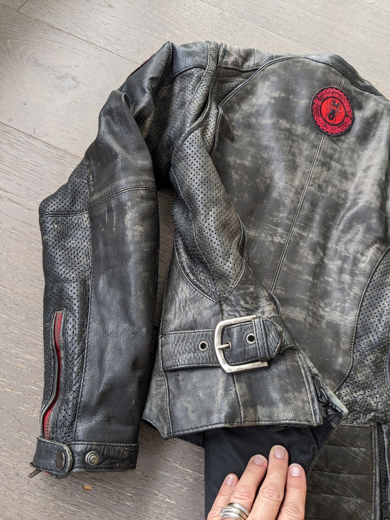 May include: Distressed black leather motorcycle jacket with a red and black patch on the back. The jacket features a buckle closure, perforated sleeves, and red zipper accents. The leather has a worn, vintage appearance.