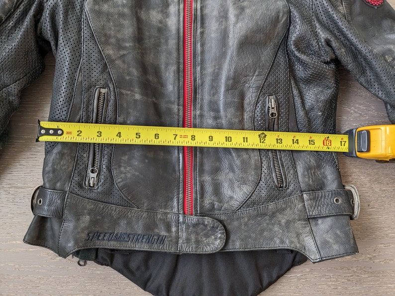 May include: A distressed black leather motorcycle jacket with a red zipper and the text "SPEED AND STRENGTH". The jacket has multiple zippered pockets and perforated panels. The jacket measures approximately 16 inches across.