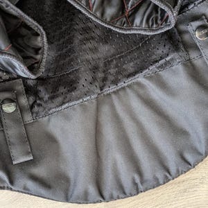 May include: Close-up of a black motorcycle jacket interior. The jacket features quilted black lining with red stitching, a breathable mesh panel, and black fabric with snap closures. The overall design suggests a focus on protection and comfort.
