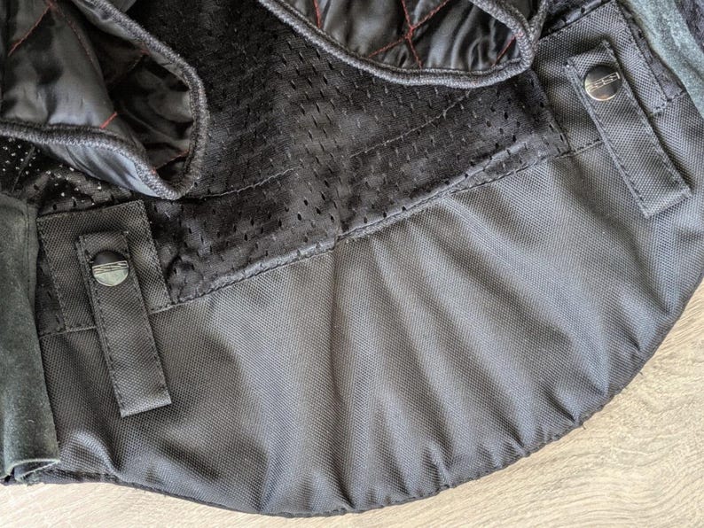 May include: Close-up of a black motorcycle jacket interior. The jacket features quilted black lining with red stitching, a breathable mesh panel, and black fabric with snap closures. The overall design suggests a focus on protection and comfort.
