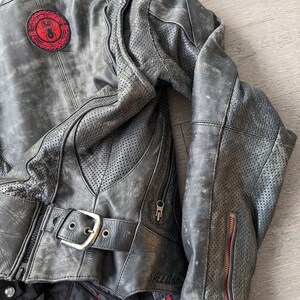 May include: Distressed black leather motorcycle jacket with a red and black patch featuring a spider. The jacket has a buckle closure, zippered pockets, and perforated panels for ventilation. The lining is black with red stitching.