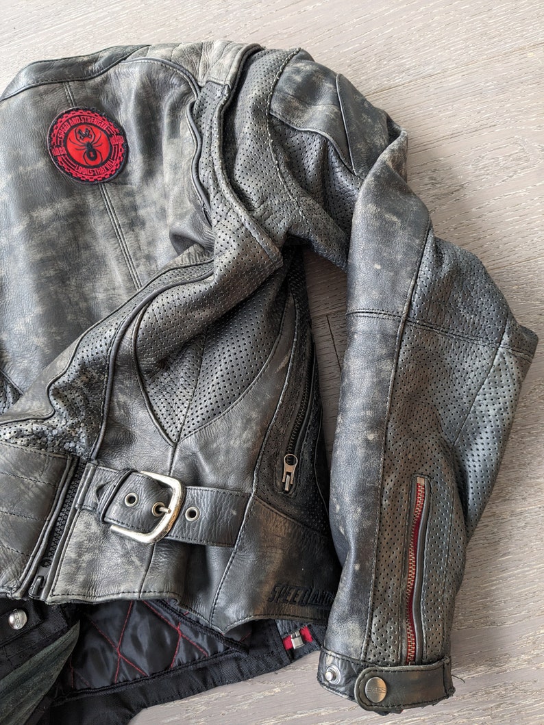 May include: Distressed black leather motorcycle jacket with a red and black patch featuring a spider. The jacket has a buckle closure, zippered pockets, and perforated panels for ventilation. The lining is black with red stitching.