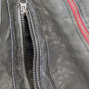 May include: Close-up of a distressed black leather jacket. The jacket features a silver zipper on the pocket and a red zipper on the seam. The leather has a worn, textured appearance with perforated details.