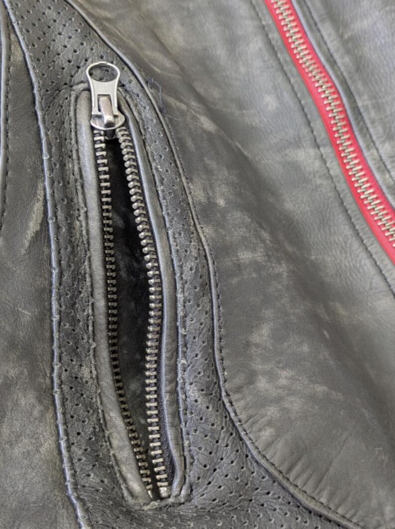 May include: Close-up of a distressed black leather jacket. The jacket features a silver zipper on the pocket and a red zipper on the seam. The leather has a worn, textured appearance with perforated details.