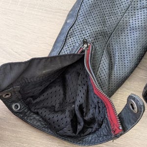 May include: Close-up of a black leather jacket sleeve with perforated detailing. The sleeve features a zippered opening with a red accent and a black mesh lining. The jacket has a textured surface and metal hardware.