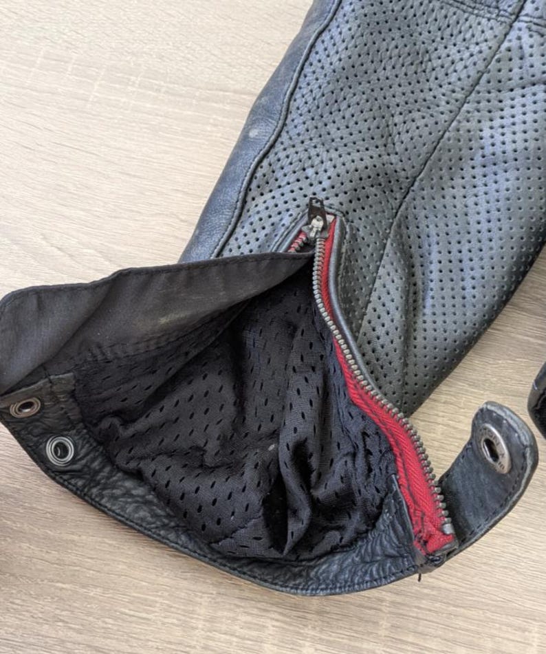 May include: Close-up of a black leather jacket sleeve with perforated detailing. The sleeve features a zippered opening with a red accent and a black mesh lining. The jacket has a textured surface and metal hardware.