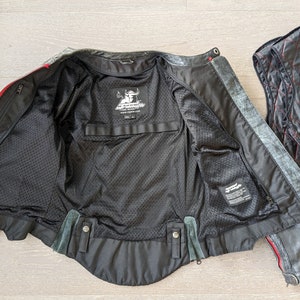 May include: Black motorcycle jacket with red accents, open to reveal a black mesh lining and interior pockets. A separate quilted vest with red stitching is next to the jacket. The jacket has a distressed finish.