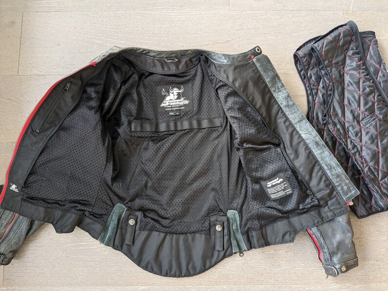 May include: Black motorcycle jacket with red accents, open to reveal a black mesh lining and interior pockets. A separate quilted vest with red stitching is next to the jacket. The jacket has a distressed finish.