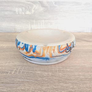 May include: A circular, off-white ceramic ashtray featuring a vibrant, swirled pattern in blue, orange, and black around its edge. The ashtray is displayed on a wooden surface.