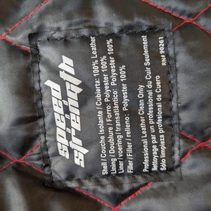 May include: Close-up of a black fabric label with the words "speed strength" in white. The label is stitched onto a black quilted fabric with red stitching. The label provides fabric composition and cleaning instructions.