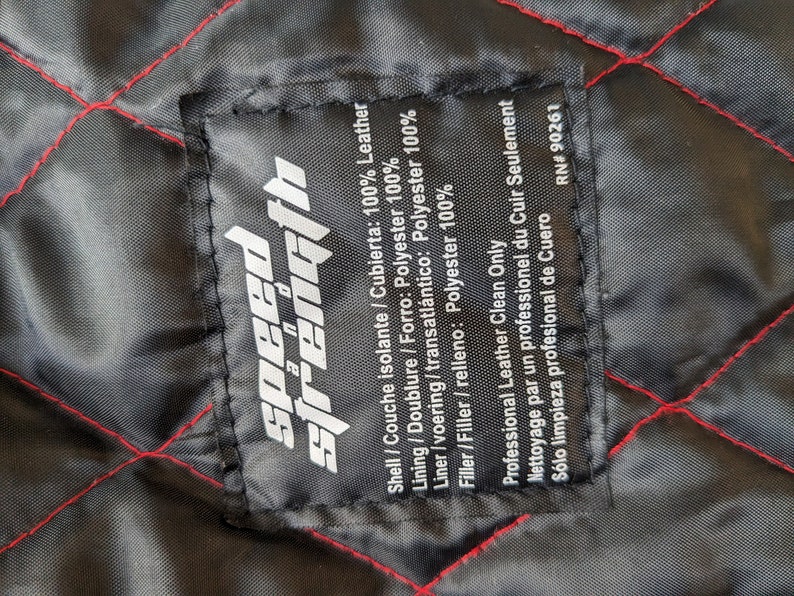 May include: Close-up of a black fabric label with the words "speed strength" in white. The label is stitched onto a black quilted fabric with red stitching. The label provides fabric composition and cleaning instructions.
