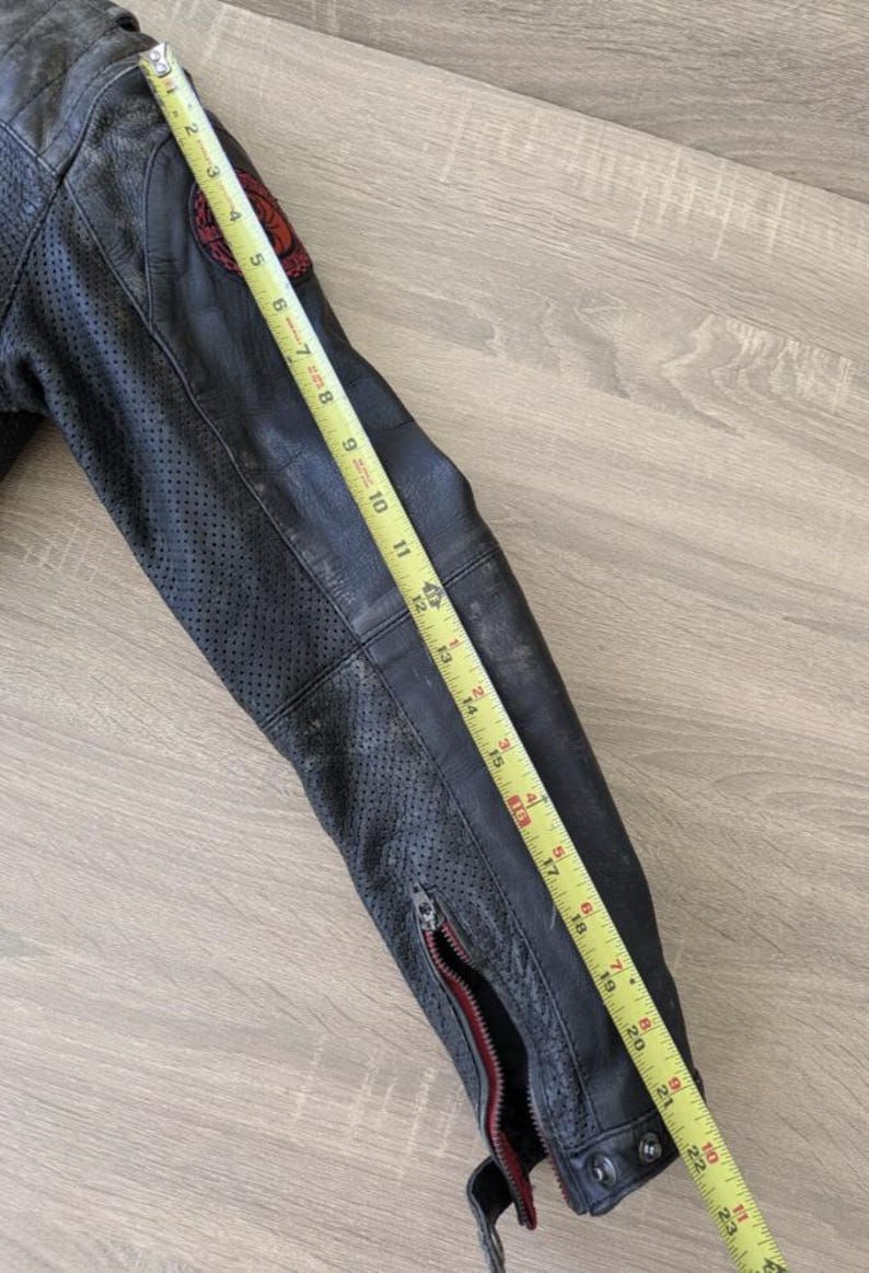 May include: Black leather motorcycle jacket sleeve with a red and black patch. The sleeve is approximately 23 inches long, featuring a zipper and perforated detailing. The jacket is worn and distressed.
