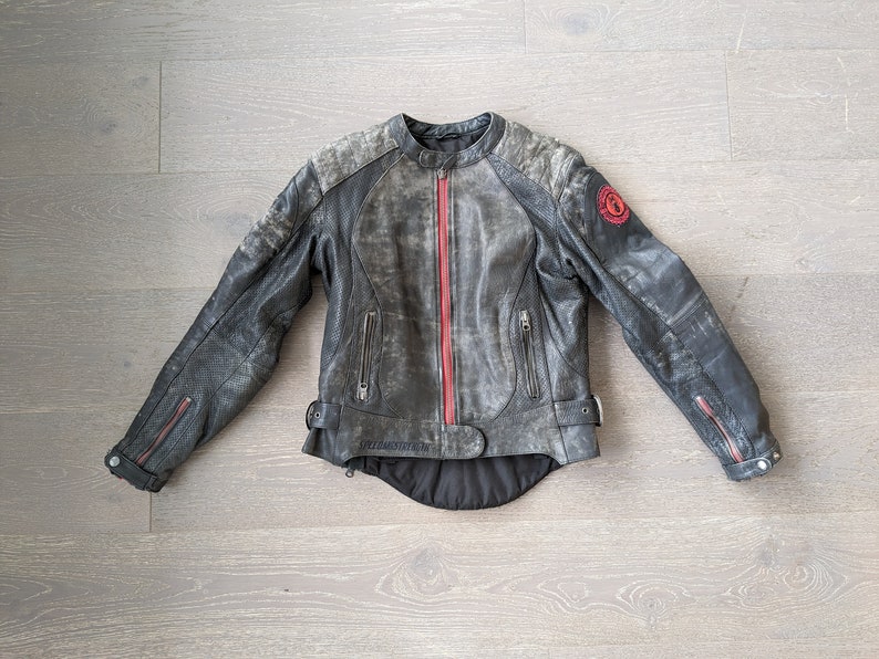 May include: A distressed gray leather motorcycle jacket with a red zipper and accents. The jacket has a round collar, zippered pockets, and a red emblem on the upper arm. The jacket is laid flat on a wooden surface.