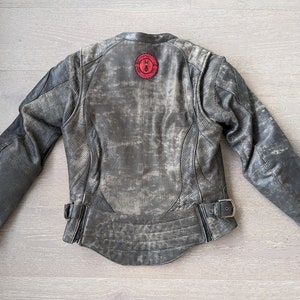 May include: Distressed gray leather motorcycle jacket with a red circular patch on the back. The jacket features perforated panels on the sleeves and a buckle closure at the waist. The jacket has a worn, vintage look.
