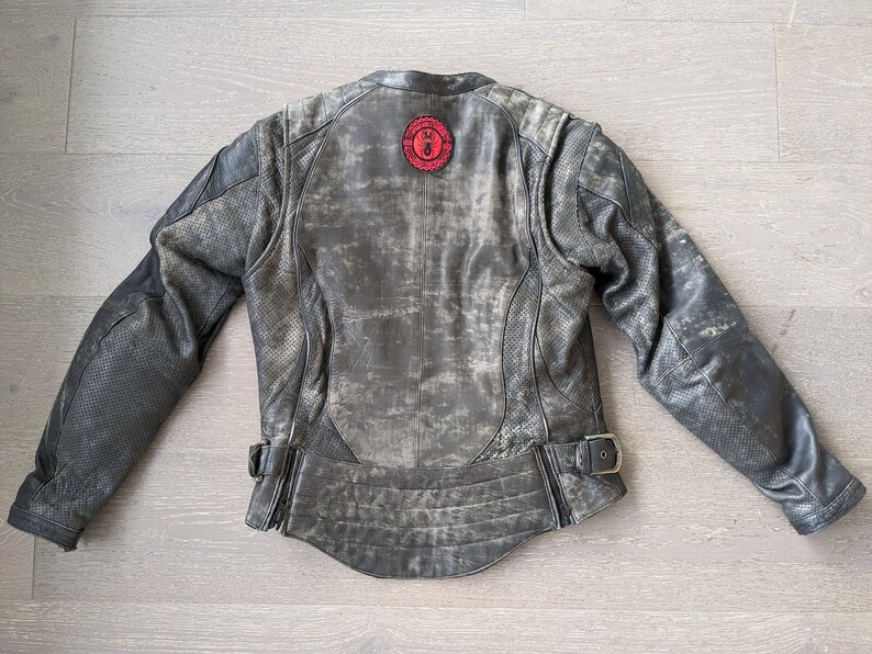 May include: Distressed gray leather motorcycle jacket with a red circular patch on the back. The jacket features perforated panels on the sleeves and a buckle closure at the waist. The jacket has a worn, vintage look.