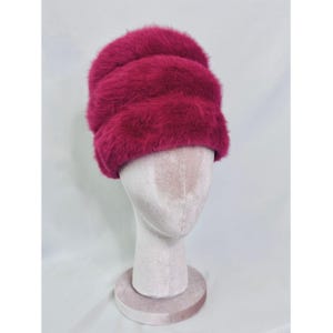 Puede incluir: Un sombrero magenta y esponjoso con un diseño acanalado horizontal. El sombrero se exhibe en una cabeza de maniquí. El sombrero parece estar hecho de un material suave y difuso, posiblemente piel o un textil similar. Es un accesorio de invierno.