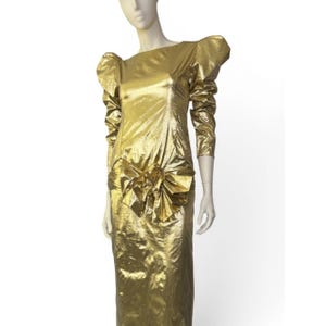 May include: A gold-coloured, long-sleeved dress with a floor-length skirt. The dress features a large, ruffled accent at the waist and puffed sleeves. The dress is displayed on a mannequin.