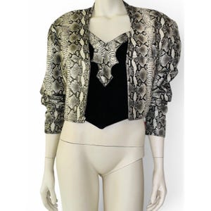 May include: A snakeskin-print jacket with a black top. The jacket has a neutral color palette of gray, white, and black. The top is black with a decorative neckline. The jacket is open in the front and has long sleeves.