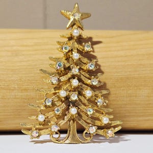 May include: A gold-tone metal Christmas tree brooch featuring a textured branch design. The tree is decorated with small white faux pearls and iridescent clear rhinestones, topped with a single star at the peak. The metal has a brushed finish.