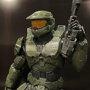 May include: A green and black action figure of a soldier wearing a helmet with a yellow visor and holding a gun.