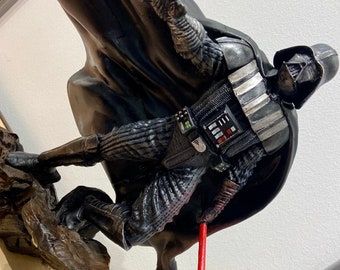 May include: A black Darth Vader action figure with a red lightsaber. The figure is posed in a dynamic action pose with a cape flowing behind it. The figure is standing on a rocky base.