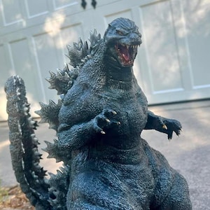May include: A close-up of a dark gray Godzilla figure with sharp teeth and claws. The figure is posed with its mouth open and its arms raised. The figure is standing on a gray surface.
