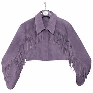 Puede incluir: Chaqueta corta de ante lavanda con mangas largas y cuello. La chaqueta presenta flecos en el pecho y a lo largo de las mangas. La chaqueta está colgada de una percha sobre un fondo blanco.