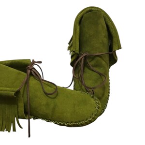 May include: Olive green suede moccasin boots with brown leather laces. The boots have a fringed top and a stitched sole. The laces are tied in a bow at the top of the foot.