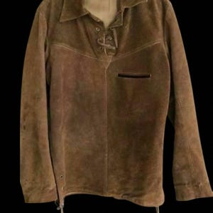 Pode incluir: Uma camisa de camurça castanha com gola e mangas compridas. A camisa apresenta um fecho de atacadores no peito e um pequeno bolso. A peça de vestuário está pendurada num cabide de plástico transparente.