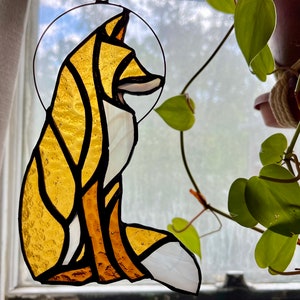 May include: A stained glass fox with a yellow and white design. The fox is sitting with its tail curled around its body. The stained glass is hanging in a window.