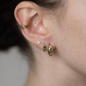 Puede incluir: Aretes de aro dorados con piedras preciosas verdes y rosas. Los aretes se muestran en la oreja de una persona con un aro de oreja dorado y un pequeño dije de ojo dorado.