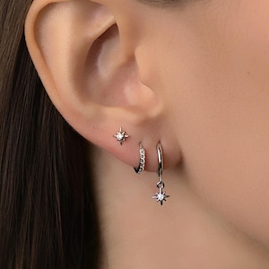 Könnte beinhalten: Nahaufnahme eines Ohrs mit drei Ohrringen. Ein kleiner sternförmiger Ohrstecker befindet sich im oberen Lappen. Ein kleiner Creolen-Ohrring mit eingebetteten Kristallen und ein hängender sternförmiger Ohrring befinden sich im unteren Lappen.