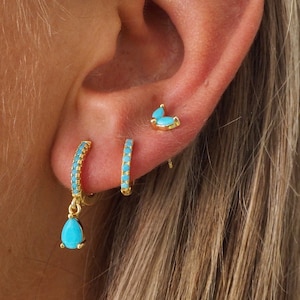 Può includere: Orecchini a cerchio dorati con pietre turchesi. Gli orecchini hanno la forma di un cerchio con un piccolo ciondolo di pietra turchese. Gli orecchini sono indossati nell'orecchio di una persona.