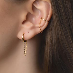 May include: Close-up of an ear with gold earrings. A small hoop earring with a chain hangs from the earlobe, and a chain cuff earring is on the upper ear. The jewellery has a delicate, elegant design.