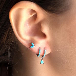 Può includere: Primo piano di un orecchio adornato da tre orecchini. L'orecchino superiore è un piccolo borchia d'argento con tre pietre color turchese. L'orecchino centrale è un piccolo cerchio d'argento con pietre turchesi. L'orecchino inferiore è un cerchio d'argento con un ciondolo a goccia turchese.