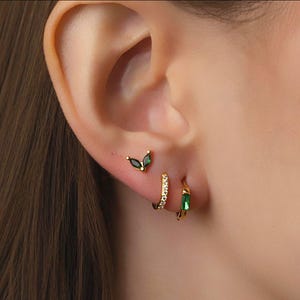 Pode incluir: Close-up de uma orelha adornada com três brincos dourados. O brinco superior é um brinco com duas pedras verdes escuras em forma de marquesa. O brinco do meio é um pequeno aro com pedras transparentes. O brinco inferior é um pequeno aro com uma pedra retangular verde.