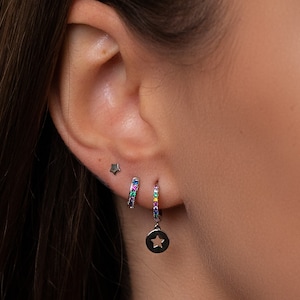 May include: A close-up of a person's ear with three silver star earrings. The earrings are a small star stud, a hoop with rainbow coloured stones, and a hoop with a dangling star charm.